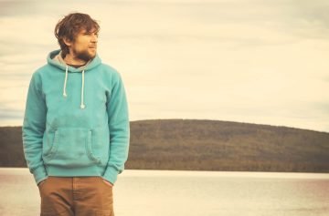 Young-Man-hipster-standing-alone-outdoor-with-mountains-and-lake-on-background-Lifestyle-and-melancholy-emotions-concept